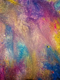 A vibrant abstract mix of colors reminiscent of powder or paint, blending seamlessly across the surface. The texture suggests grains or small clumps intermingling with flowing hues.