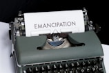 A vintage typewriter with a dark green body has a sheet of paper loaded that reads 'EMANCIPATION' in large bold letters. The background is mostly black, providing a stark contrast to the white paper.