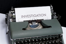 A vintage typewriter with a page inserted, featuring the word 'INVESTIGATION' typed at the top. The typewriter is dark green with black keys and metal components, set against a contrasting black and white background.