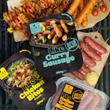 Various packaged plant-based meat products labeled as gluten-free and organic are displayed alongside cooked sausages and skewers of vegetables. The packages include items like 'Like Curry Sausage', 'Like Chicken Bites', and 'Like Italian Griller'. Bright, fresh ingredients such as cherry tomatoes and mustard are artfully arranged on a grill and wood cutting boards.