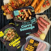 Various packaged plant-based meat products labeled as gluten-free and organic are displayed alongside cooked sausages and skewers of vegetables. The packages include items like 'Like Curry Sausage', 'Like Chicken Bites', and 'Like Italian Griller'. Bright, fresh ingredients such as cherry tomatoes and mustard are artfully arranged on a grill and wood cutting boards.