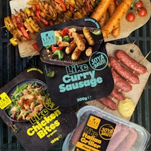 Various packaged plant-based meat products labeled as gluten-free and organic are displayed alongside cooked sausages and skewers of vegetables. The packages include items like 'Like Curry Sausage', 'Like Chicken Bites', and 'Like Italian Griller'. Bright, fresh ingredients such as cherry tomatoes and mustard are artfully arranged on a grill and wood cutting boards.