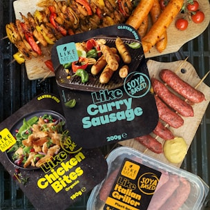 Various packaged plant-based meat products labeled as gluten-free and organic are displayed alongside cooked sausages and skewers of vegetables. The packages include items like 'Like Curry Sausage', 'Like Chicken Bites', and 'Like Italian Griller'. Bright, fresh ingredients such as cherry tomatoes and mustard are artfully arranged on a grill and wood cutting boards.