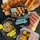 Various packaged plant-based meat products labeled as gluten-free and organic are displayed alongside cooked sausages and skewers of vegetables. The packages include items like 'Like Curry Sausage', 'Like Chicken Bites', and 'Like Italian Griller'. Bright, fresh ingredients such as cherry tomatoes and mustard are artfully arranged on a grill and wood cutting boards.