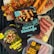 Various packaged plant-based meat products labeled as gluten-free and organic are displayed alongside cooked sausages and skewers of vegetables. The packages include items like 'Like Curry Sausage', 'Like Chicken Bites', and 'Like Italian Griller'. Bright, fresh ingredients such as cherry tomatoes and mustard are artfully arranged on a grill and wood cutting boards.