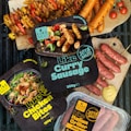 Various packaged plant-based meat products labeled as gluten-free and organic are displayed alongside cooked sausages and skewers of vegetables. The packages include items like 'Like Curry Sausage', 'Like Chicken Bites', and 'Like Italian Griller'. Bright, fresh ingredients such as cherry tomatoes and mustard are artfully arranged on a grill and wood cutting boards.
