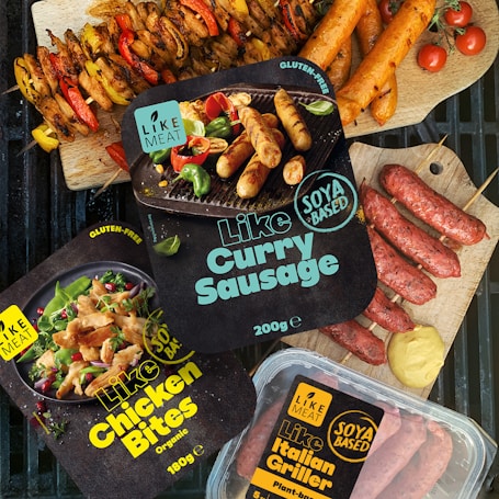 Various packaged plant-based meat products labeled as gluten-free and organic are displayed alongside cooked sausages and skewers of vegetables. The packages include items like 'Like Curry Sausage', 'Like Chicken Bites', and 'Like Italian Griller'. Bright, fresh ingredients such as cherry tomatoes and mustard are artfully arranged on a grill and wood cutting boards.