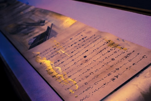 An aged piece of paper with handwritten text in a flowing script is laid out, complemented by splashes of gold paint. The text appears to be a poem or a letter. The lighting casts a gentle glow, creating shadows that highlight the texture of the paper.