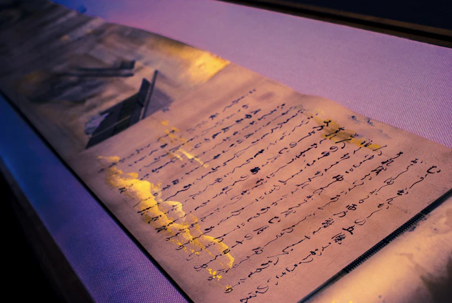 An aged piece of paper with handwritten text in a flowing script is laid out, complemented by splashes of gold paint. The text appears to be a poem or a letter. The lighting casts a gentle glow, creating shadows that highlight the texture of the paper.