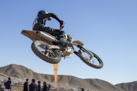 A motorcyclist performs an airborne stunt on a dirt bike against a clear blue sky. The rider wears black protective gear and a helmet, while spectators observe from below, with hills in the background.
