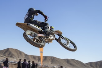 A motorcyclist performs an airborne stunt on a dirt bike against a clear blue sky. The rider wears black protective gear and a helmet, while spectators observe from below, with hills in the background.