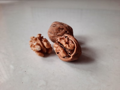 Two whole walnuts and a walnut half are placed on a smooth surface with a slightly reflective quality. The nuts' intricate textures and deep brown color stand out against the light gray background, creating a natural and earthy aesthetic.