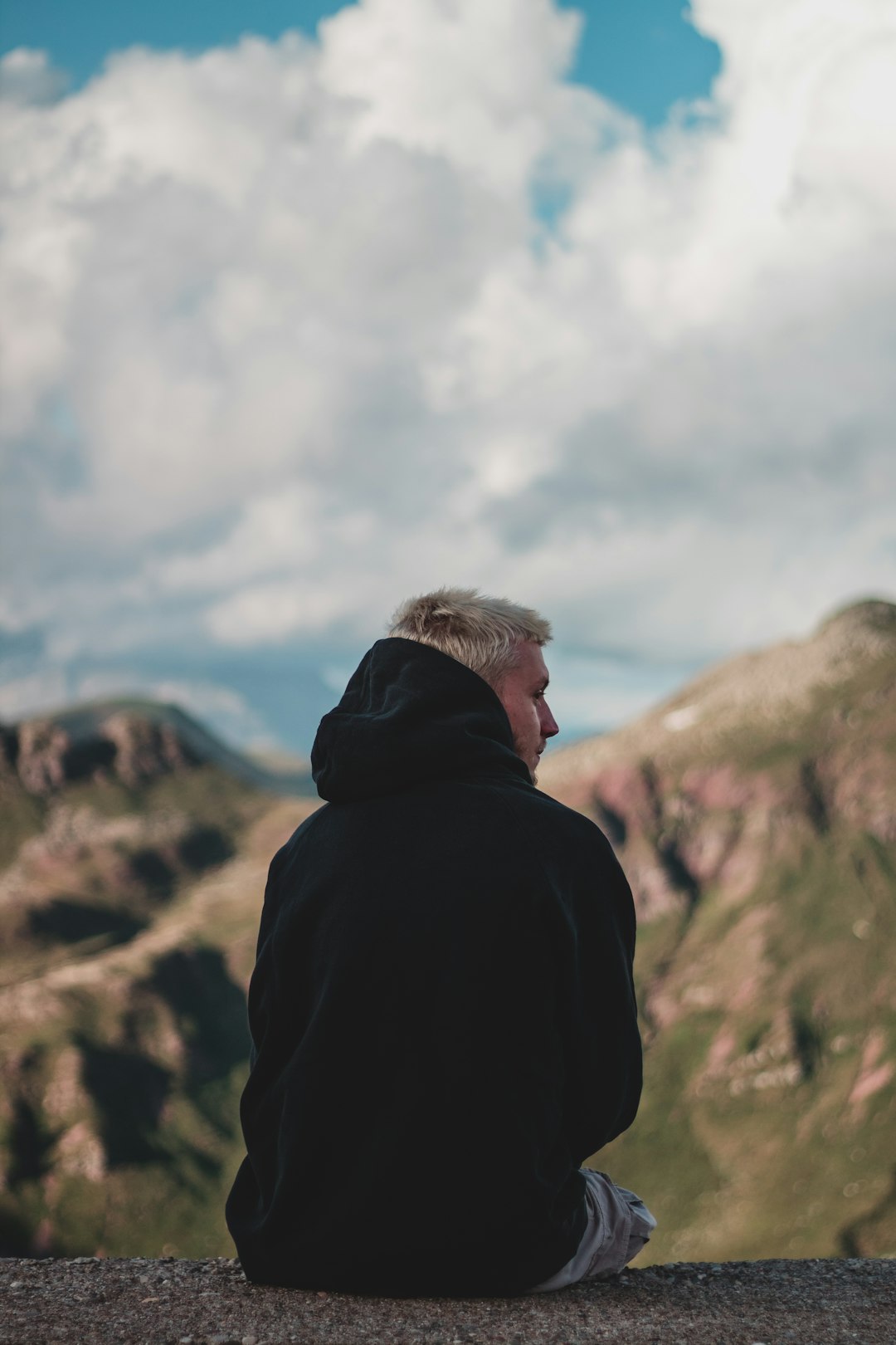 man in black hoodie standing on brown rock mountain under white clouds during daytime