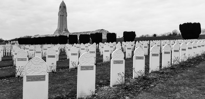 A cemetery with rows of elaborately designed white tombstones, each with inscriptions, set in a grassy area. In the background, a tall stone monument or tower stands, surrounded by trimmed hedges. The overcast sky adds to the somber ambiance of the scene.