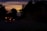 An ambulance with its lights on is traveling down a dimly lit road during sunset. The sky displays a gradient of warm colors, including orange and purple, as the sun sets beyond the horizon. Trees line the road, creating a silhouette effect against the colorful sky.