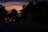 An ambulance with its lights on is traveling down a dimly lit road during sunset. The sky displays a gradient of warm colors, including orange and purple, as the sun sets beyond the horizon. Trees line the road, creating a silhouette effect against the colorful sky.