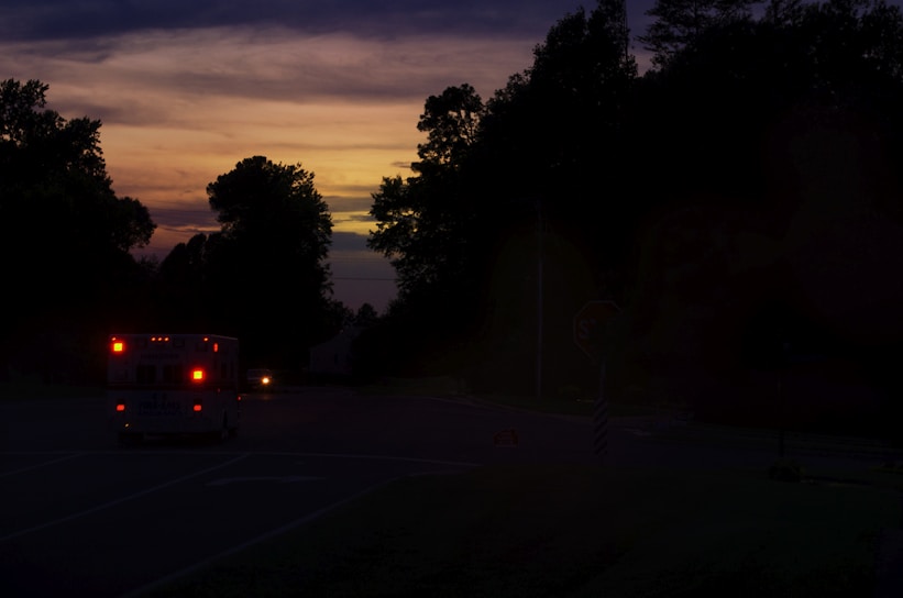 An ambulance with its lights on is traveling down a dimly lit road during sunset. The sky displays a gradient of warm colors, including orange and purple, as the sun sets beyond the horizon. Trees line the road, creating a silhouette effect against the colorful sky.