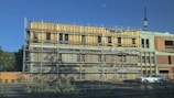 A mid-construction building with scaffolding surrounding it, featuring multiple levels. The structure appears to be made of a combination of unfinished wood and concrete. A car is parked on the nearby road, and a large crane is visible in the background. Vegetation is present in the foreground, and the sky is clear and blue.