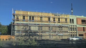 A mid-construction building with scaffolding surrounding it, featuring multiple levels. The structure appears to be made of a combination of unfinished wood and concrete. A car is parked on the nearby road, and a large crane is visible in the background. Vegetation is present in the foreground, and the sky is clear and blue.