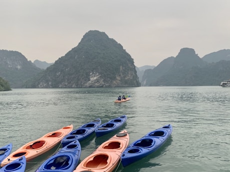 Several kayaks are floating on calm water with a backdrop of large, lush green mountains under an overcast sky. Two people in a kayak are paddling towards the mountains.