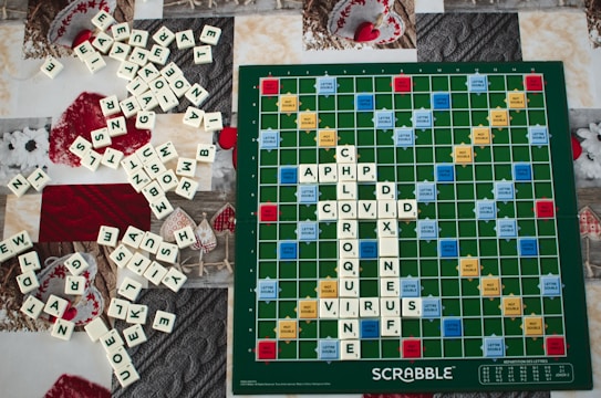 A Scrabble board with several words spelled out using white letter tiles, including 'CHLOROQUINE' and 'COVID' intersecting. A large number of additional tiles are scattered around the board on a textured surface, featuring a variety of patterns and holiday decorations in red and white.