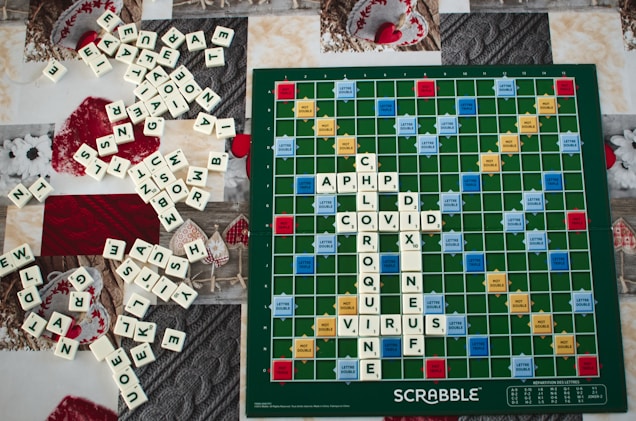A Scrabble board with several words spelled out using white letter tiles, including 'CHLOROQUINE' and 'COVID' intersecting. A large number of additional tiles are scattered around the board on a textured surface, featuring a variety of patterns and holiday decorations in red and white.