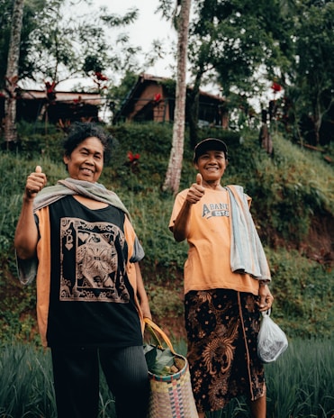 Two people are standing outdoors surrounded by lush greenery and trees. They are smiling and giving a thumbs-up gesture. One is holding a woven basket with vegetables, and the other has a plastic bag. Both are dressed in casual clothing, and small houses are visible in the background nestled amongst the trees.