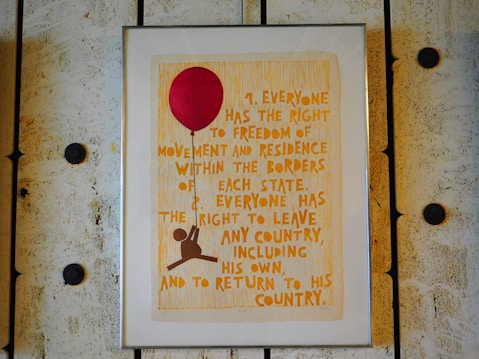 A framed poster with text that outlines the right to freedom of movement and residence within national borders. The poster is adorned with a large red balloon and an illustration of a figure holding onto the balloon string. The frame and wall have a rustic, textured appearance.