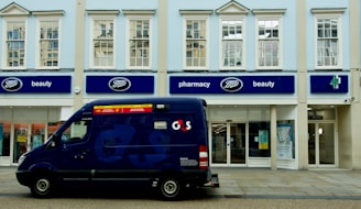 A blue security van is parked in front of a store with signage for Boots, a retail chain known for pharmacy and beauty products. The building is painted in light colors, with large windows on the upper floor. The storefront features blue signs and the Boots logo.