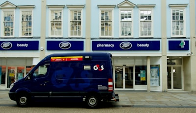 A blue security van is parked in front of a store with signage for Boots, a retail chain known for pharmacy and beauty products. The building is painted in light colors, with large windows on the upper floor. The storefront features blue signs and the Boots logo.
