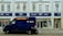 A blue security van is parked in front of a store with signage for Boots, a retail chain known for pharmacy and beauty products. The building is painted in light colors, with large windows on the upper floor. The storefront features blue signs and the Boots logo.
