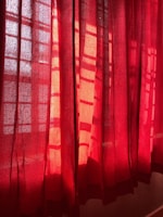 Red curtains partially drawn, allowing sunlight to filter through, creating patterns of light and shadow on the fabric. The material has a slightly translucent quality, revealing a hint of the scene beyond.