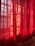 Red curtains partially drawn, allowing sunlight to filter through, creating patterns of light and shadow on the fabric. The material has a slightly translucent quality, revealing a hint of the scene beyond.