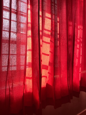 Red curtains partially drawn, allowing sunlight to filter through, creating patterns of light and shadow on the fabric. The material has a slightly translucent quality, revealing a hint of the scene beyond.