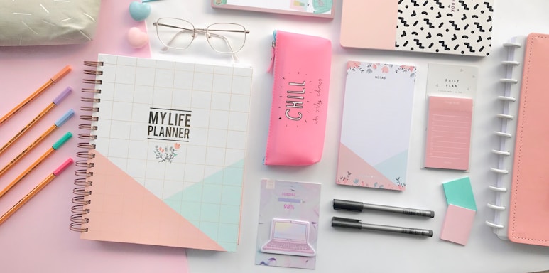 A collection of stationery items arranged neatly on a pastel-colored surface. The items include a spiral-bound planner with a geometric design, a pink pencil case with the word 'CHILL', several pastel-colored pens and pencils, notepads, glasses, and other accessories. The colors are soft and calming, predominantly pink, white, and mint green.
