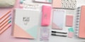 A collection of stationery items arranged neatly on a pastel-colored surface. The items include a spiral-bound planner with a geometric design, a pink pencil case with the word 'CHILL', several pastel-colored pens and pencils, notepads, glasses, and other accessories. The colors are soft and calming, predominantly pink, white, and mint green.
