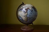 A globe is placed on a wooden table against a light green wall. The globe features detailed geographical markings, showing various countries in Africa and Asia, with the Indian Ocean prominent. The stand of the globe is simple and wooden, complementing the dark metal axis that supports the globe.