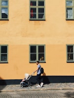 A person is pushing a stroller along a cobblestone street next to a yellow building with multiple green-trimmed windows. The sunlight creates sharp shadows, and the scene is calm and picturesque.