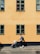 A person is pushing a stroller along a cobblestone street next to a yellow building with multiple green-trimmed windows. The sunlight creates sharp shadows, and the scene is calm and picturesque.