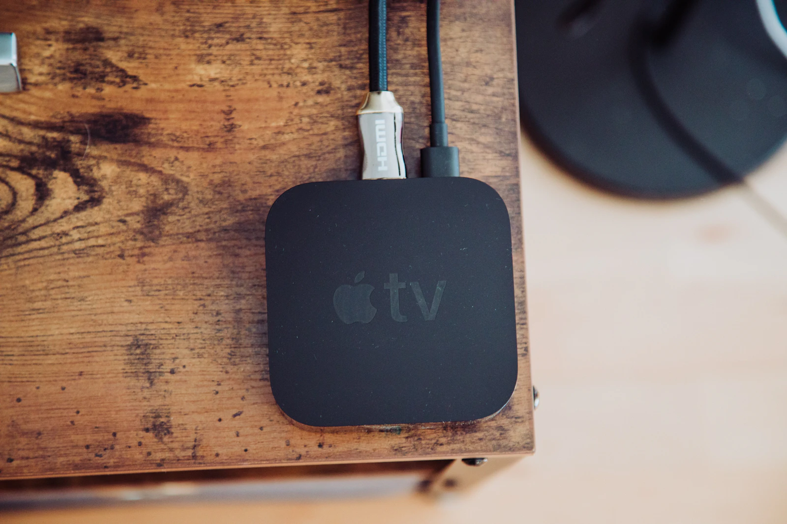 Black Apple TV streaming box on a rustic wooden shelf with a silver HDMI cable and a black power cord plugged into the back