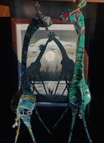 Two wire and metal sculptures portray giraffes with intertwined necks, set against a framed painting that features the silhouettes of giraffes reaching towards a full moon amidst the landscape. The composition is enhanced by vibrant colors on the sculptures and muted greys and blacks in the painting.