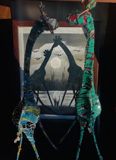 Two wire and metal sculptures portray giraffes with intertwined necks, set against a framed painting that features the silhouettes of giraffes reaching towards a full moon amidst the landscape. The composition is enhanced by vibrant colors on the sculptures and muted greys and blacks in the painting.