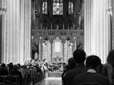 A large and ornate cathedral interior featuring high vaulted ceilings and intricate stained glass windows. Several rows of seated people are visible, facing towards an altar or stage area where a group of musicians appears to be performing.