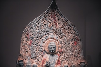 An intricate stone carving featuring a central figure resembling Buddha, surrounded by a detailed halo and ornate patterns. The figure is flanked by smaller carved figures, with an overall design suggesting religious or spiritual significance. The artwork displays a weathered texture, adding to its historical atmosphere.