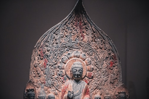 An intricate stone carving featuring a central figure resembling Buddha, surrounded by a detailed halo and ornate patterns. The figure is flanked by smaller carved figures, with an overall design suggesting religious or spiritual significance. The artwork displays a weathered texture, adding to its historical atmosphere.