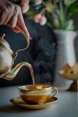 A hand is elegantly pouring tea from a brass teapot into a matching brass teacup, with visible steam rising from the cup. The background features a soft-focus image of a vase with colorful flowers.