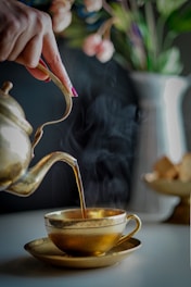 A hand is elegantly pouring tea from a brass teapot into a matching brass teacup, with visible steam rising from the cup. The background features a soft-focus image of a vase with colorful flowers.
