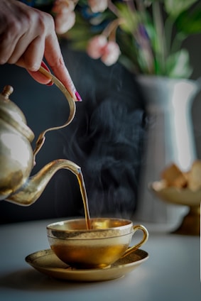 A hand is elegantly pouring tea from a brass teapot into a matching brass teacup, with visible steam rising from the cup. The background features a soft-focus image of a vase with colorful flowers.