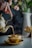 A hand is elegantly pouring tea from a brass teapot into a matching brass teacup, with visible steam rising from the cup. The background features a soft-focus image of a vase with colorful flowers.
