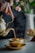 A hand is elegantly pouring tea from a brass teapot into a matching brass teacup, with visible steam rising from the cup. The background features a soft-focus image of a vase with colorful flowers.